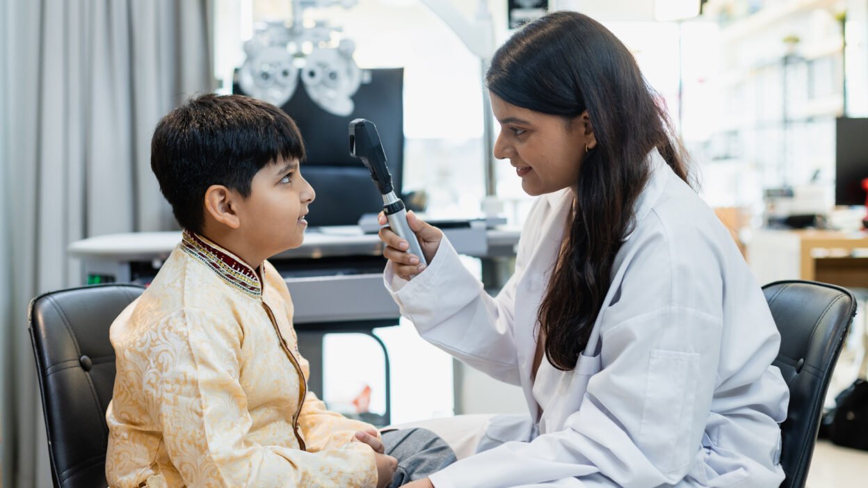 Indian child boy with women optometrist being examined for eyesight follow up using a modern optometry equipment called a retinoscope in clinic.