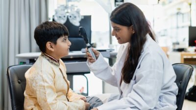 Indian child boy with women optometrist being examined for eyesight follow up using a modern optometry equipment called a retinoscope in clinic.